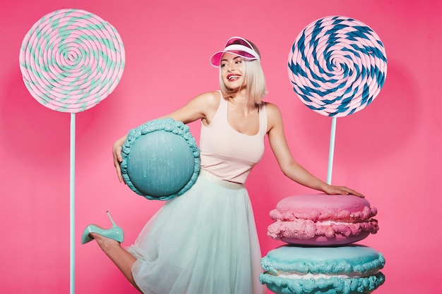 Młoda dziewczyna z blond włosami na sobie top i spódnicę stojącą z ogromnym słodkim lizakiem i makaronikami na różowo