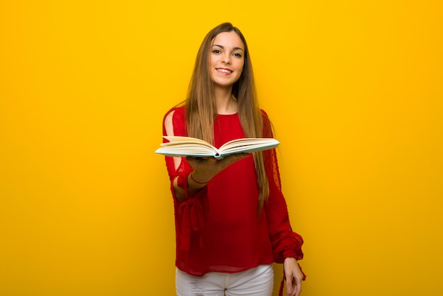 Młoda dziewczyna z czerwoną sukienkę na żółtej ścianie, trzymając książkę i dając to komuś