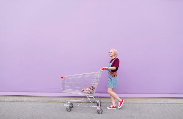 młoda dziewczyna z wózek na zakupy idąc ulicą na tle fioletowej ściany