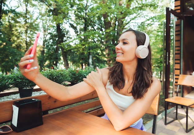 Młoda dziewczyna ze słuchawkami pije letniego orzeźwiającego drinka w miejskiej kawiarni i robi sobie selfie