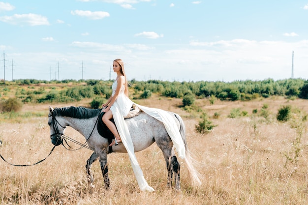 Młoda elegancka dziewczyna w długiej białej sukni flowy pozowanie na koniu na wsi