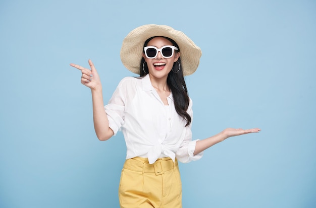 Młoda elegancka piękna Azjatycka kobieta ubrana w letnie ubrania, uśmiechając się i wskazując na pustą przestrzeń kopii na białym tle na niebieskim tle.