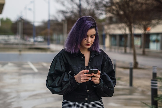 Młoda Europejka z fioletowymi włosami spacerująca po ulicy i wysyłająca SMS-y na swoim telefonie