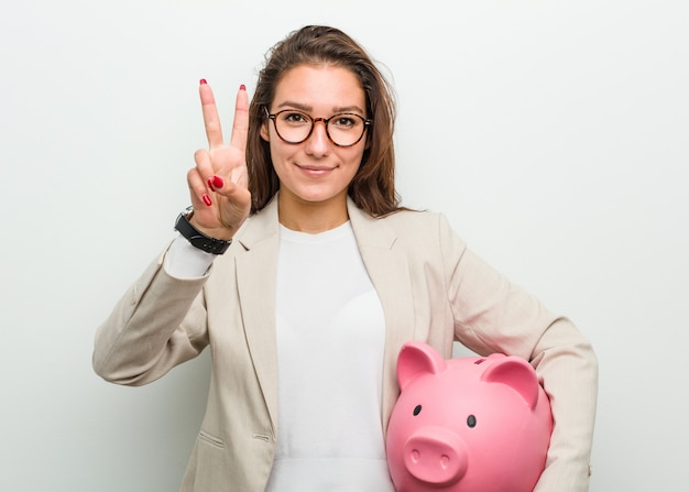 Młoda europejska biznesowa kobieta trzyma prosiątko banka pokazuje numer dwa z palcami.