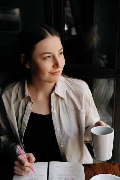 Młoda europejska freelancerka o długich ciemnych włosach pije kawę i pracuje zdalnie w kawiarni