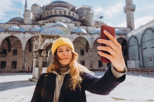 Młoda Europejska Kobieta Bierze Selfie Portret W Istanbuł, Turcja. Dziewczyna Idzie Przez Zimowy Stambuł. Blondynka Robi Zdjęcie Telefonem Na Tle Meczetu W Jesienny Dzień.