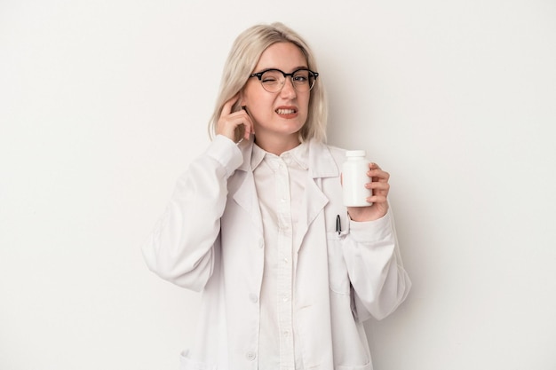 Młoda farmaceuta kobieta trzymająca tabletki na białym tle zakrywająca uszy rękami