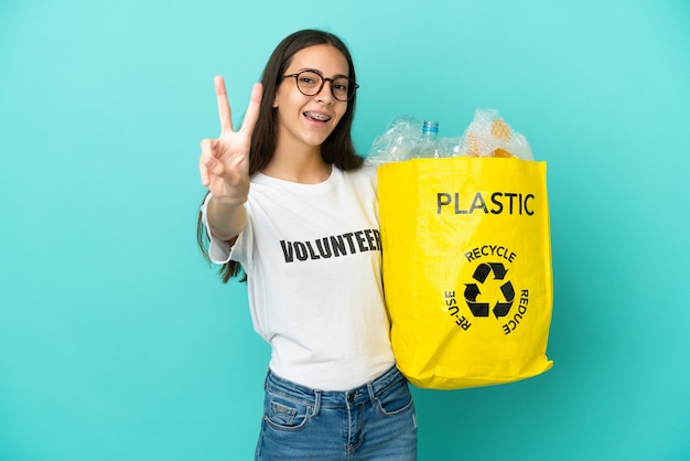 Młoda Francuzka trzyma torbę pełną plastikowych butelek do recyklingu, uśmiechając się i pokazując znak zwycięstwa