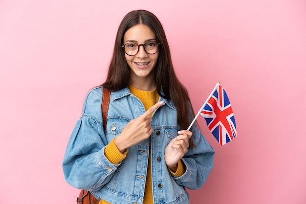 Młoda Francuzka trzymająca flagę Wielkiej Brytanii odizolowaną na różowym tle skierowaną w bok, aby zaprezentować produkt