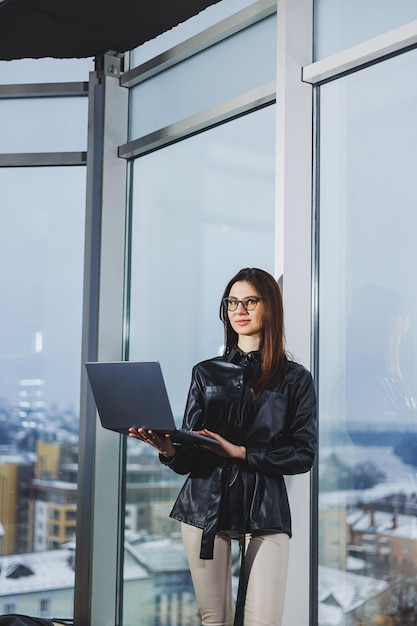 Młoda freelancer kobieta w okularach z laptopem pracująca zdalnie w nowoczesnym obszarze roboczym z dużymi oknami Praca zdalna