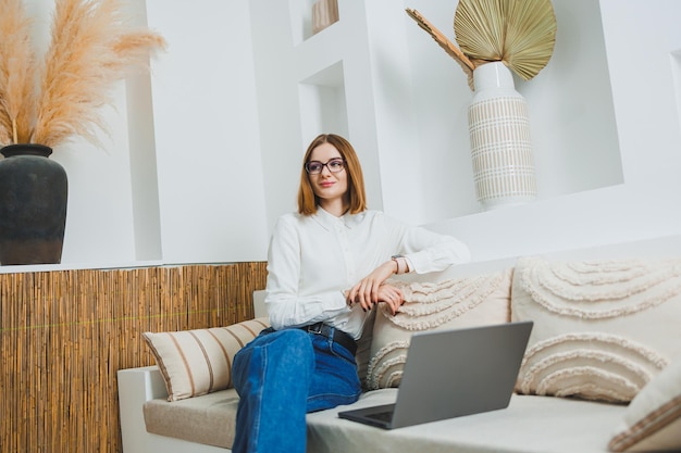 Zdjęcie młoda freelancerka siedząca na kanapie i pracująca na laptopie oglądająca film na laptopie studiująca blogowanie relaksująca się i rozmawiająca online