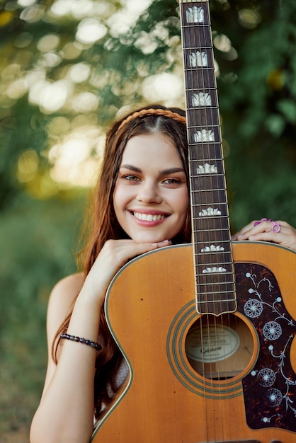Zdjęcie młoda hipis kobieta z eko wizerunkiem uśmiecha się i patrzy w kamerę z gitarą w ręku w naturze podczas podróży