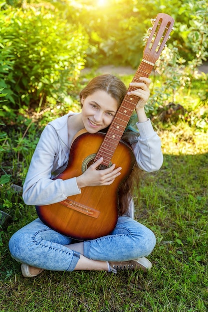 Młoda hipster kobieta siedzi w trawie i gra na gitarze na tle parku lub ogrodu Nastoletnia dziewczyna uczy się grać piosenki i pisać muzykę