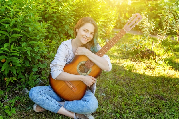 Młoda hipster kobieta siedzi w trawie i gra na gitarze na tle parku lub ogrodu Nastoletnia dziewczyna uczy się grać piosenki i pisać muzykę