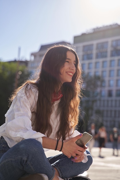 Młoda Hipster Kobieta Z Długimi Włosami Trzymająca Inteligentne Urządzenie Telefoniczne Za Pomocą Aplikacji Mobilnej Siedząca Na Zewnątrz