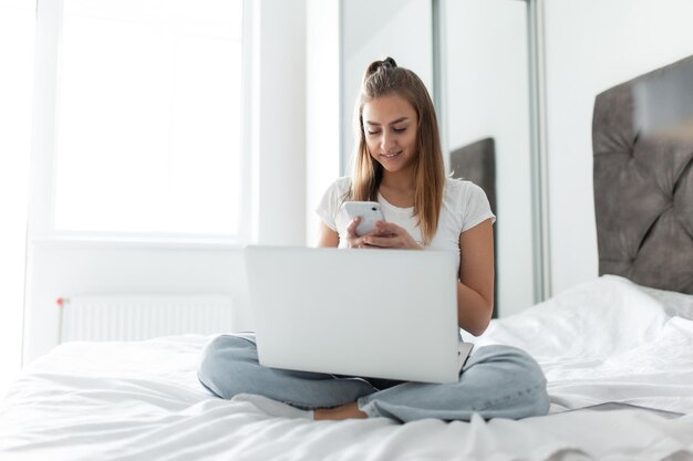 Młoda hipsterka używająca smartfona i laptopa, siedząc na łóżku w jasnym pokoju, nowoczesne gadżety