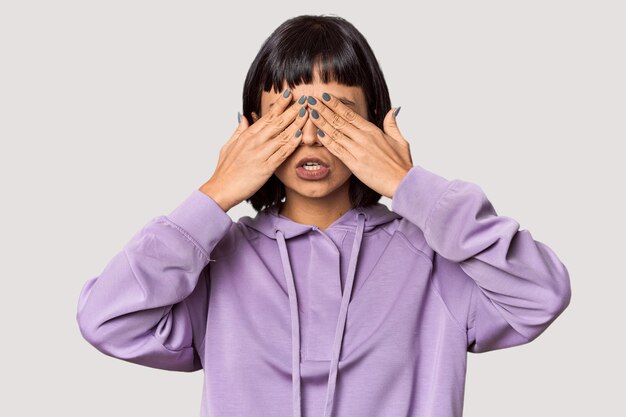 Młoda hiszpańska kobieta z krótkimi czarnymi włosami w studiu boi się zakrywać oczy rękami