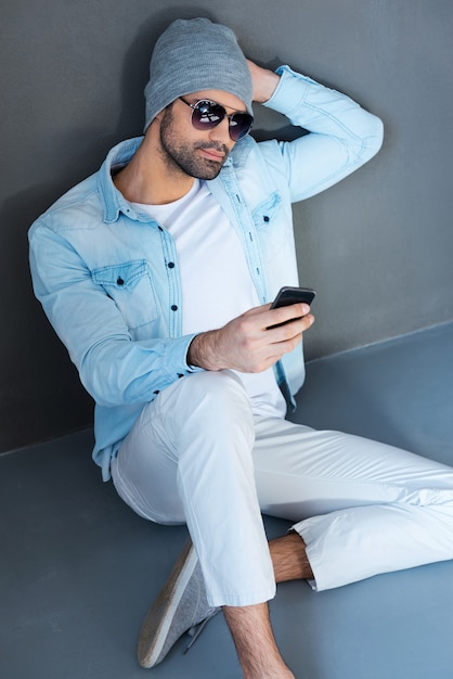 Młoda i beztroska. Widok z góry przystojnego młodego mężczyzny w okularach trzymającego telefon komórkowy, siedząc na podłodze na szarym tle