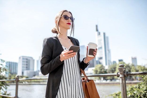 Młoda i elegancka bizneswoman spacerująca z telefonem i kawą na nabrzeżu z wieżowcami we Frankfurcie
