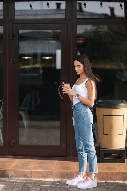 Młoda i piękna kobieta używa telefonu na zewnątrz przed kawiarnią Atrakcyjna brunetka w białej koszuli