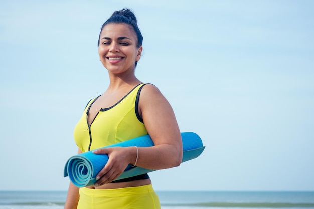 Młoda indyjska kobieta ćwiczy asany na niebieskiej macie do jogi na plaży Goa, ubrana w stylowe żółte legginsy i stanik fitness