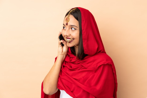 Młoda indyjska kobieta na beżowej ścianie prowadząca z kimś rozmowę z telefonem komórkowym
