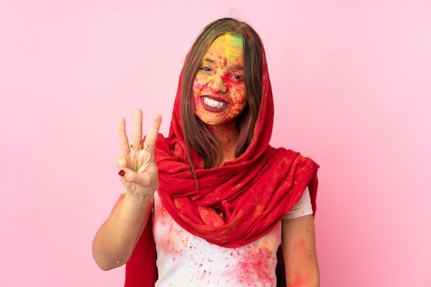 Młoda indyjska kobieta z kolorowymi holi pudrami na twarzy na różowej ścianie szczęśliwa i licząca trzy z palcami