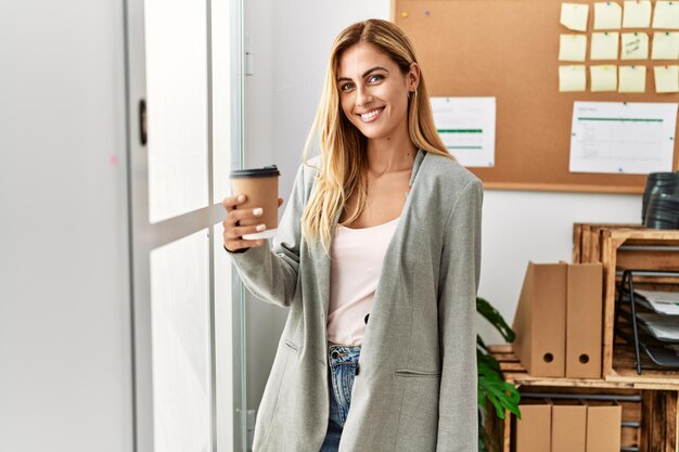 Młoda kaukaska bizneswoman pijąca kawę stojącą przy oknie w biurze