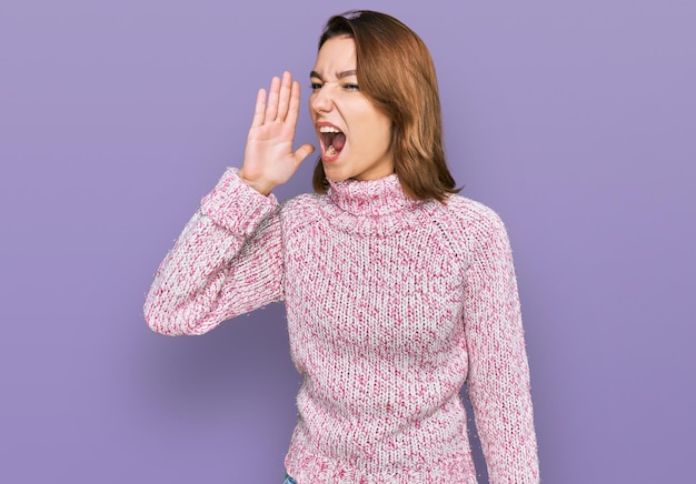 Młoda kaukaska dziewczyna ubrana w wełniany zimowy sweter krzyczy i krzyczy głośno na bok z ręką na ustach koncepcja komunikacji