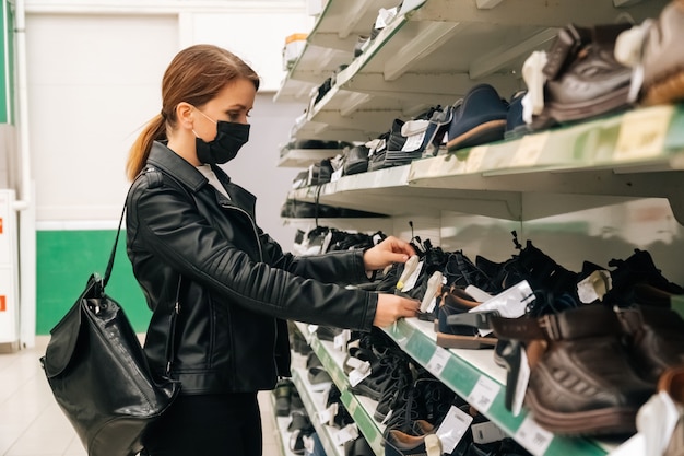 Zdjęcie młoda kaukaska dziewczyna w czarnej medycznej masce wybiera ubrania, buty w supermarkecie. pojęcie dystansu społecznego i