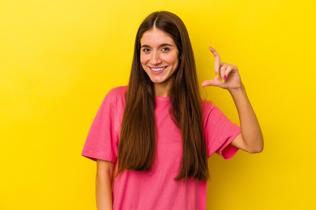 Młoda kaukaska kobieta na białym tle na żółtym tle trzymająca coś małego palcami wskazującymi, uśmiechnięta i pewna siebie.