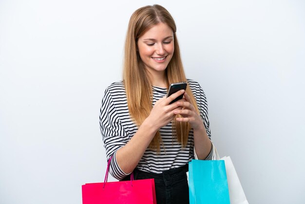 Młoda kaukaska kobieta na białym tle trzymająca torby na zakupy i pisząca wiadomość telefonem komórkowym do przyjaciela