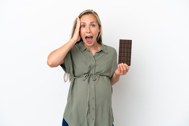 Młoda kaukaska kobieta na białym tle w ciąży i trzymająca czekoladę z zaskoczonym wyrazem twarzy