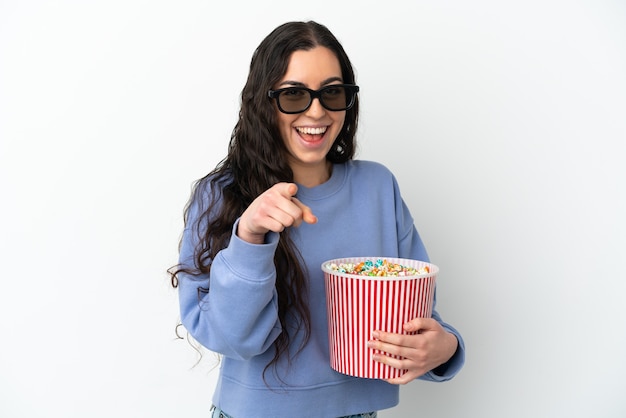 Młoda kaukaska kobieta na białym tle w okularach 3d i trzymająca duże wiadro popcornów, wskazując przód