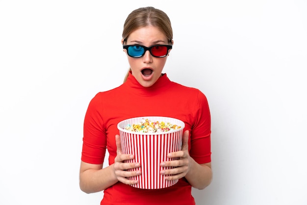 Młoda kaukaska kobieta na białym tle zaskoczona okularami 3d i trzyma duże wiadro popcorns