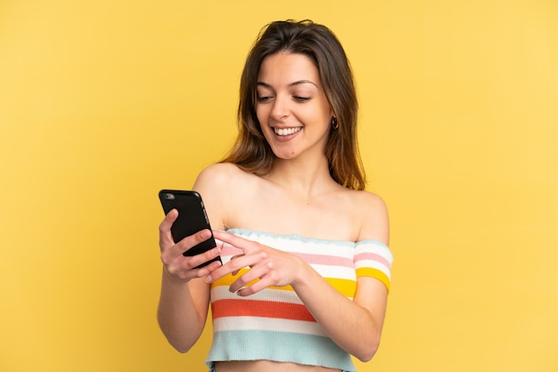 Młoda kaukaska kobieta na żółtym tle wysyła wiadomość lub e-mail za pomocą telefonu komórkowego