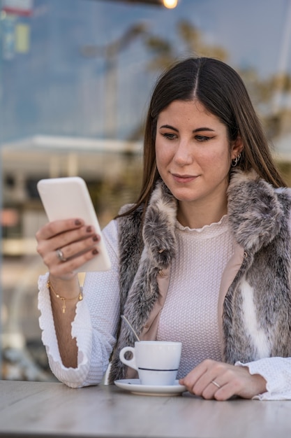 Młoda kaukaska kobieta pije kawę, trzymając telefon komórkowy