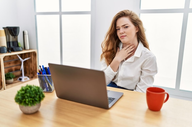 Młoda kaukaska kobieta pracująca w biurze za pomocą laptopa dotykająca bolesnej szyi, bólu gardła na grypę, grudkę i infekcję