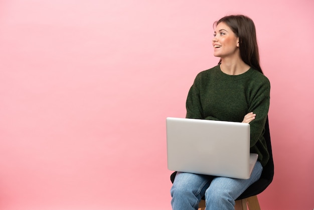 Młoda kaukaska kobieta siedzi na krześle z laptopem na białym tle na różowym tle szczęśliwa i uśmiechnięta