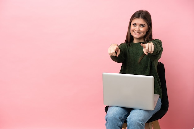 Młoda kaukaska kobieta siedzi na krześle z laptopem na białym tle na różowym tle, wskazując palcem na ciebie, uśmiechając się