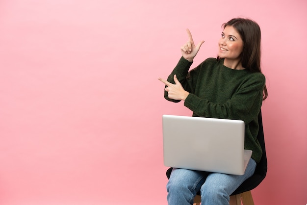 Młoda kaukaska kobieta siedzi na krześle z laptopem na białym tle, wskazując palcem wskazującym, to świetny pomysł