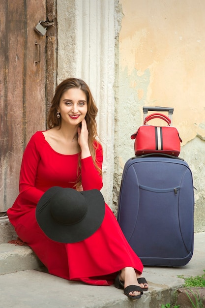 Młoda kaukaska kobieta siedzi na progu starego domu w czerwonej długiej sukience z walizką