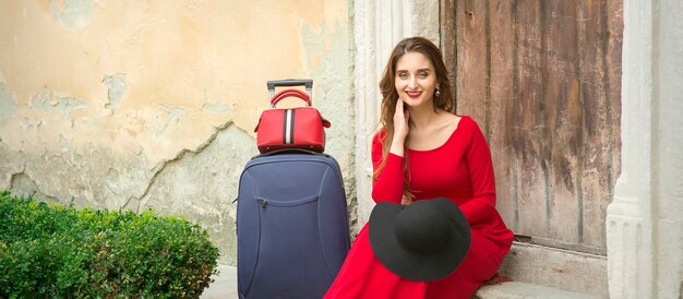 Młoda kaukaska kobieta siedzi na progu starego domu w czerwonej długiej sukience z walizką