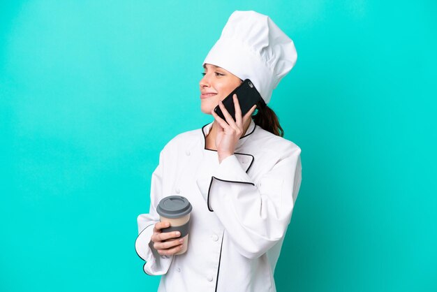 Młoda kaukaska kobieta szefa kuchni na białym tle na niebieskim tle trzyma kawę na wynos i telefon komórkowy