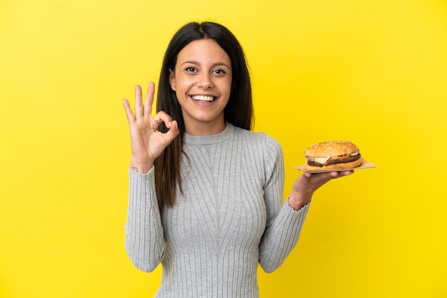 Młoda kaukaska kobieta trzyma burgera na białym tle na żółtym tle pokazując znak ok palcami