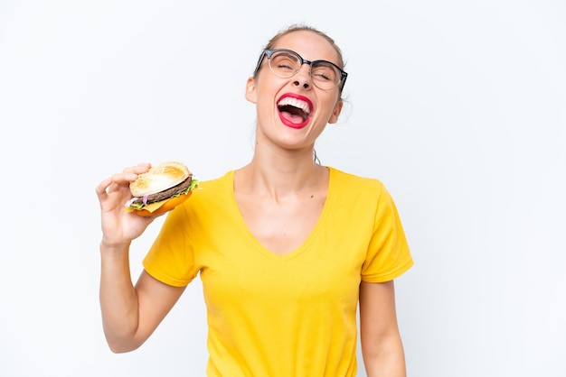 Młoda kaukaska kobieta trzyma hamburgera odizolowywającego na białym tle śmieje się