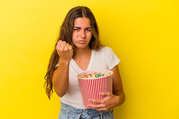 Młoda kaukaska kobieta trzyma popcorns na białym tle na żółtym tle pokazując pięść do kamery, agresywny wyraz twarzy.