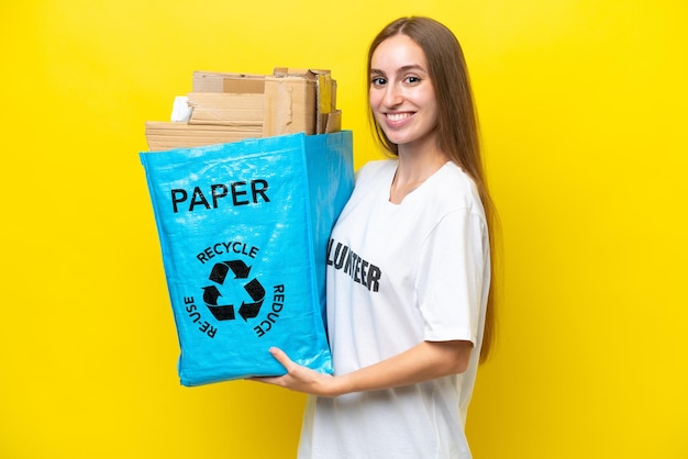 Młoda kaukaska kobieta trzyma torbę do recyklingu pełną papieru do recyklingu odizolowaną na żółtym tle, uśmiechając się dużo