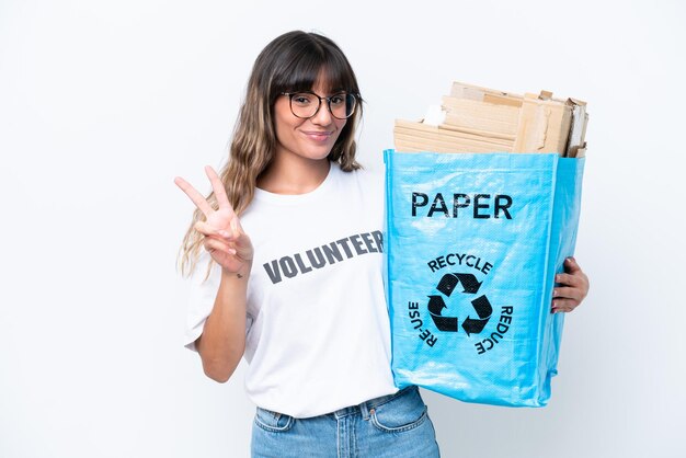 Młoda kaukaska kobieta trzyma worek do recyklingu pełen papieru do recyklingu na białym tle, uśmiechając się i pokazując znak zwycięstwa
