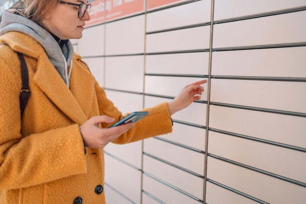 Młoda kaukaska kobieta trzymająca smartfon w pobliżu automatycznego terminala pocztowego inteligentna dostawa samoobsługowa po